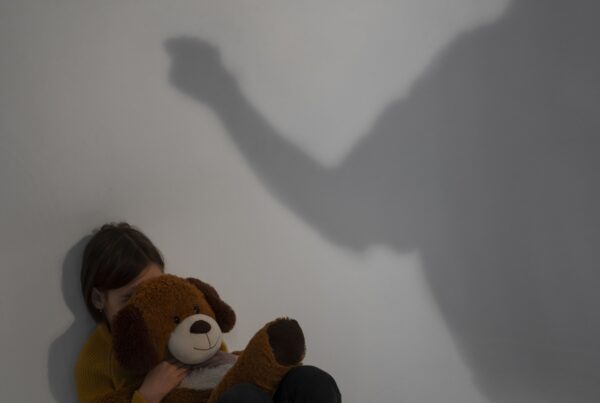 Bambino impaurito che abbraccia un peluche mentre un’ombra adulta minacciosa alza il braccio: rappresentazione simbolica di violenza domestica e pregiudizio per il minore.