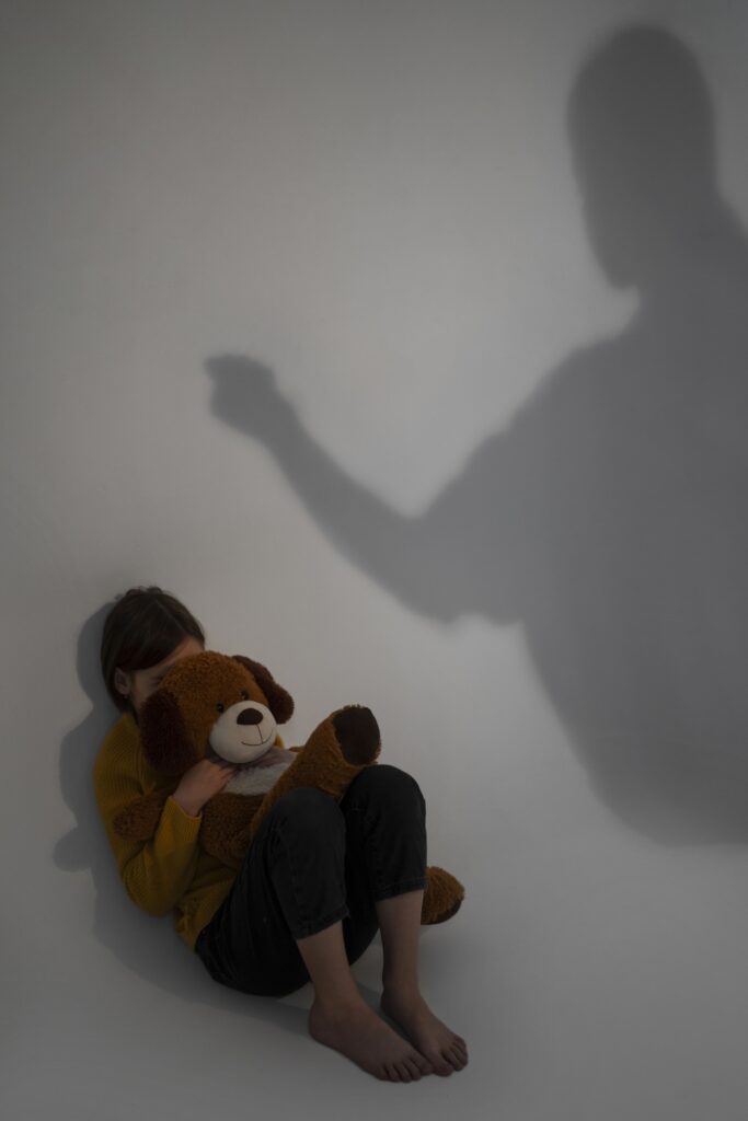 Bambino impaurito che abbraccia un peluche mentre un’ombra adulta minacciosa alza il braccio: rappresentazione simbolica di violenza domestica e pregiudizio per il minore.